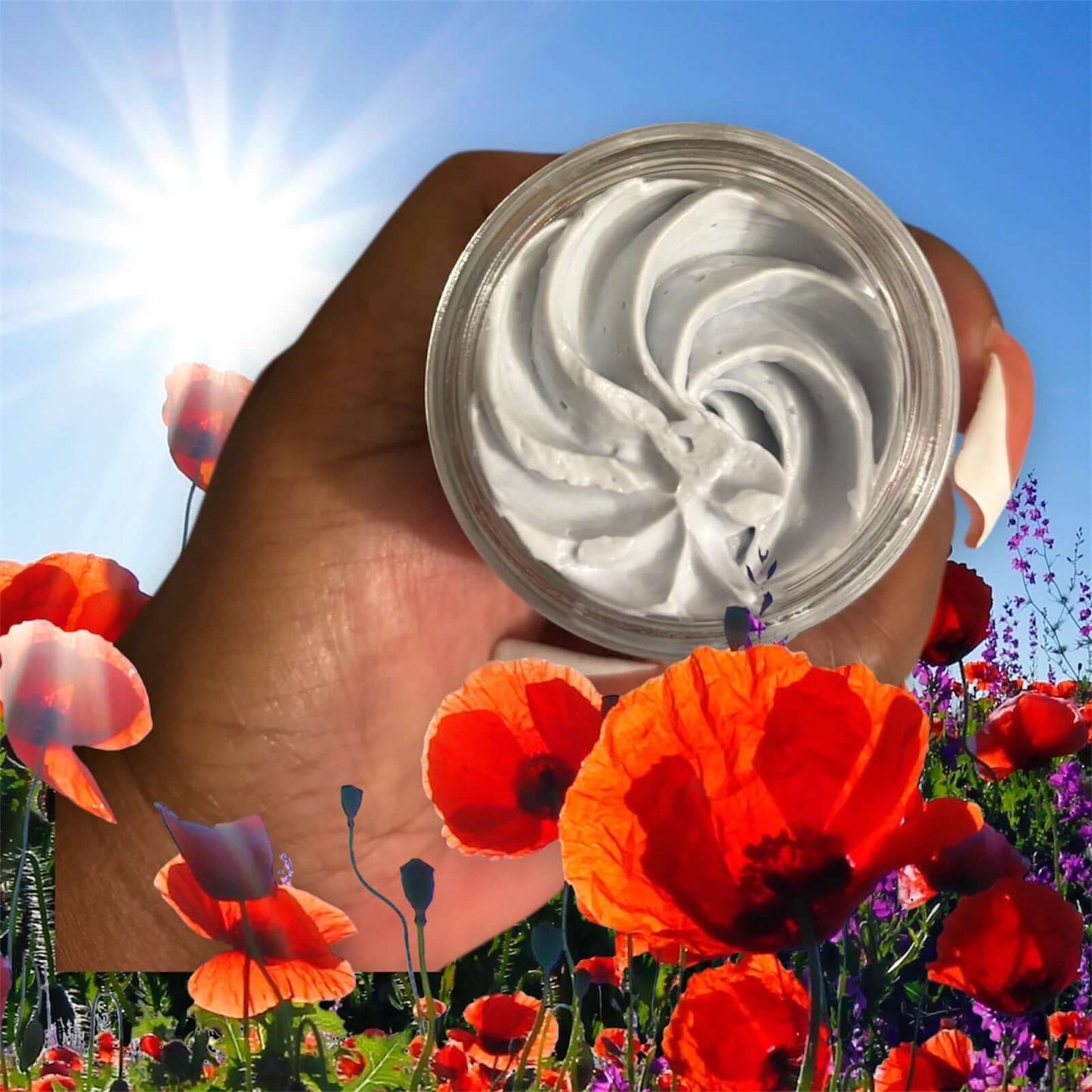 Organic whipped body butter in a clear jar, surrounded by vibrant red flowers and bright sunlight.
